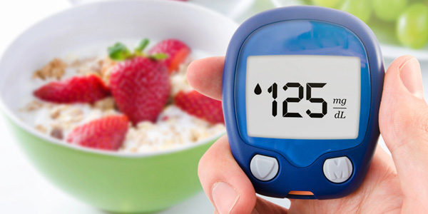 Hand hält ein blaues Blutzuckermessgerät mit Anzeige „125 mg/dL“. Im Hintergrund eine Schale mit Müsli, Erdbeeren, weiteren Früchten und grüne Weintrauben. Motiv zeigt visuell eine Blutzuckermessung im Ernährungskontext.